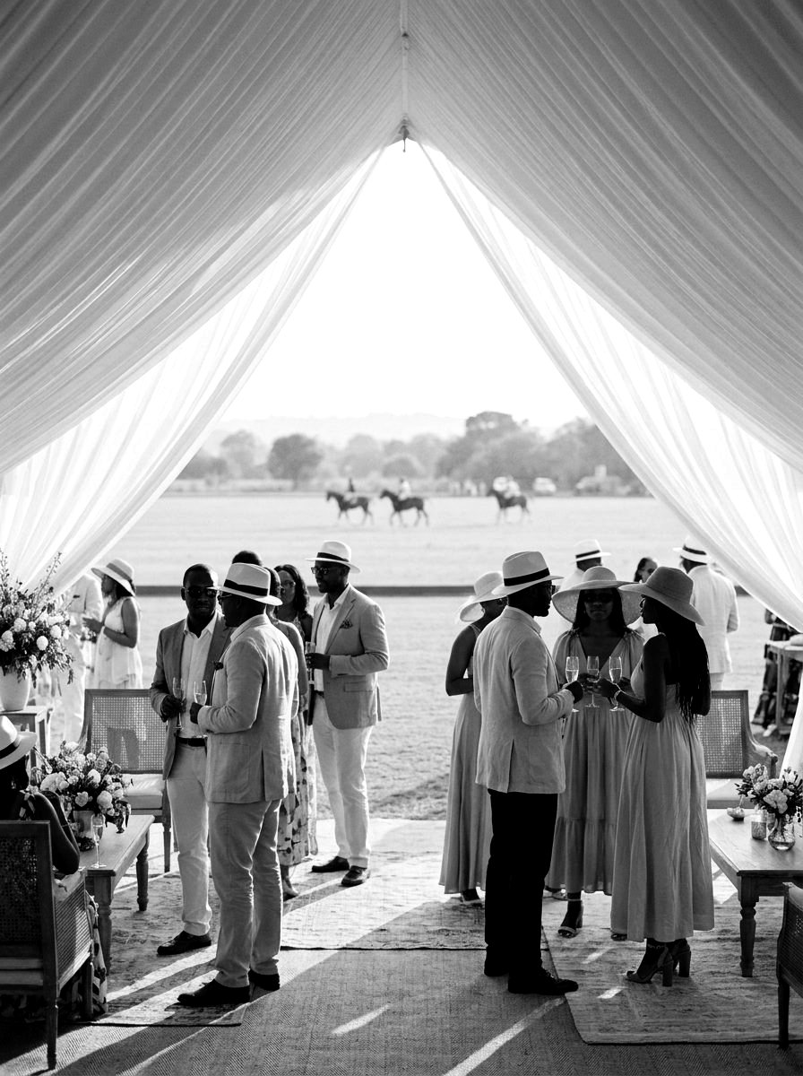 Guests in summer linen inside a draped marquee at the edge of a polo field